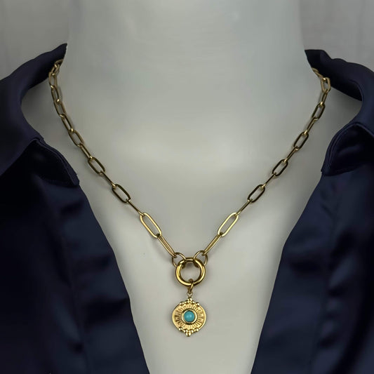 A gold paperclip chain necklace with a circular ring connector and a round gold pendant featuring a turquoise blue stone, displayed on a white mannequin bust wearing a dark navy satin shirt.