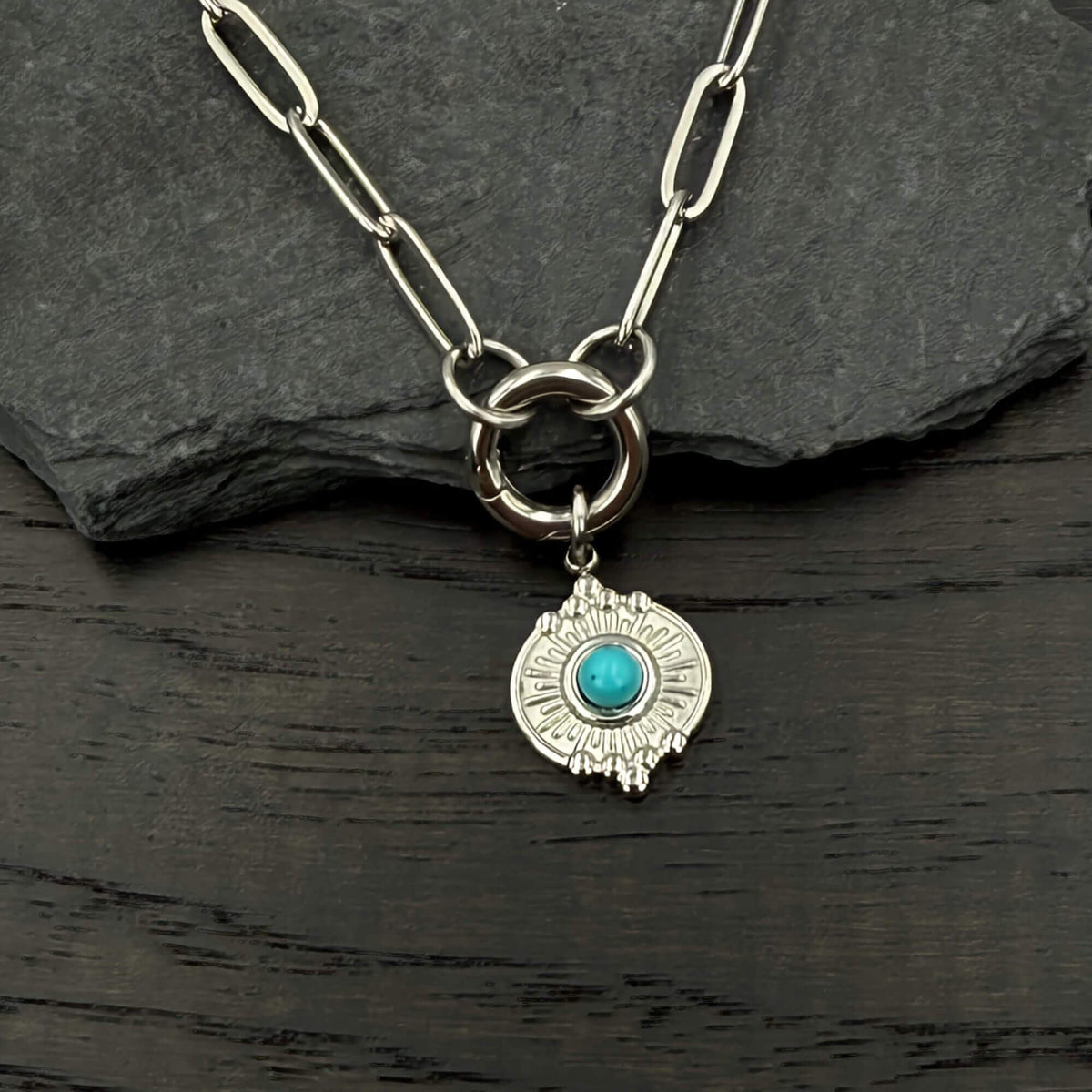 A silver-toned chain necklace with a circular ring clasp and a dangling shield-shaped charm featuring a turquoise stone, displayed on a dark slate and wood surface.