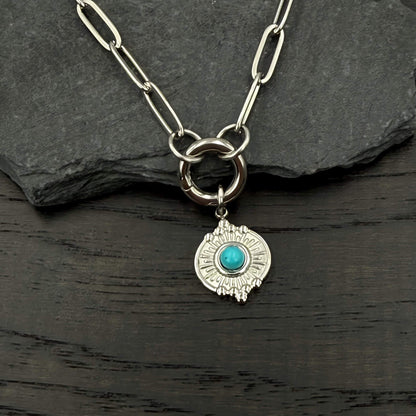 A silver-toned chain necklace with a circular ring clasp and a dangling shield-shaped charm featuring a turquoise stone, displayed on a dark slate and wood surface.