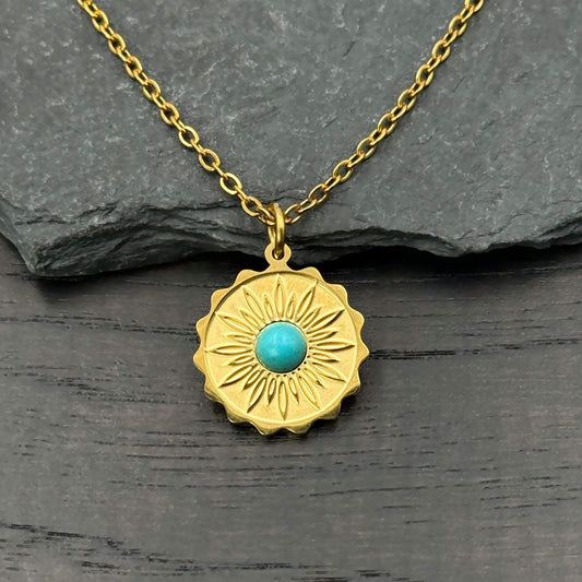 A gold-toned sunburst pendant necklace with a central turquoise blue stone, displayed on a dark gray slate surface.