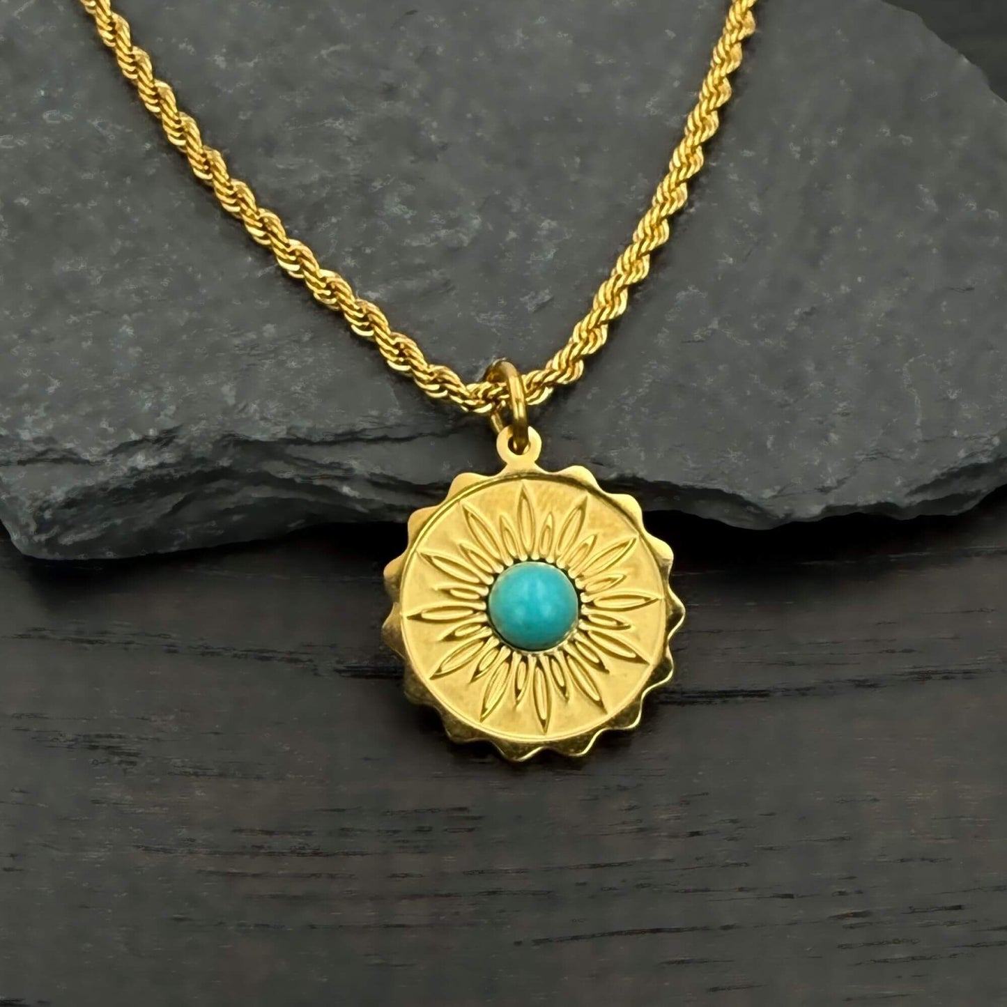 A gold-toned necklace with a textured sunburst pendant featuring a central turquoise stone, displayed on a dark slate surface.