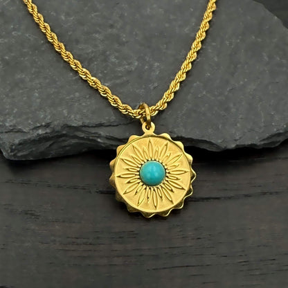 A gold-toned necklace with a textured sunburst pendant featuring a central turquoise stone, displayed on a dark slate surface.