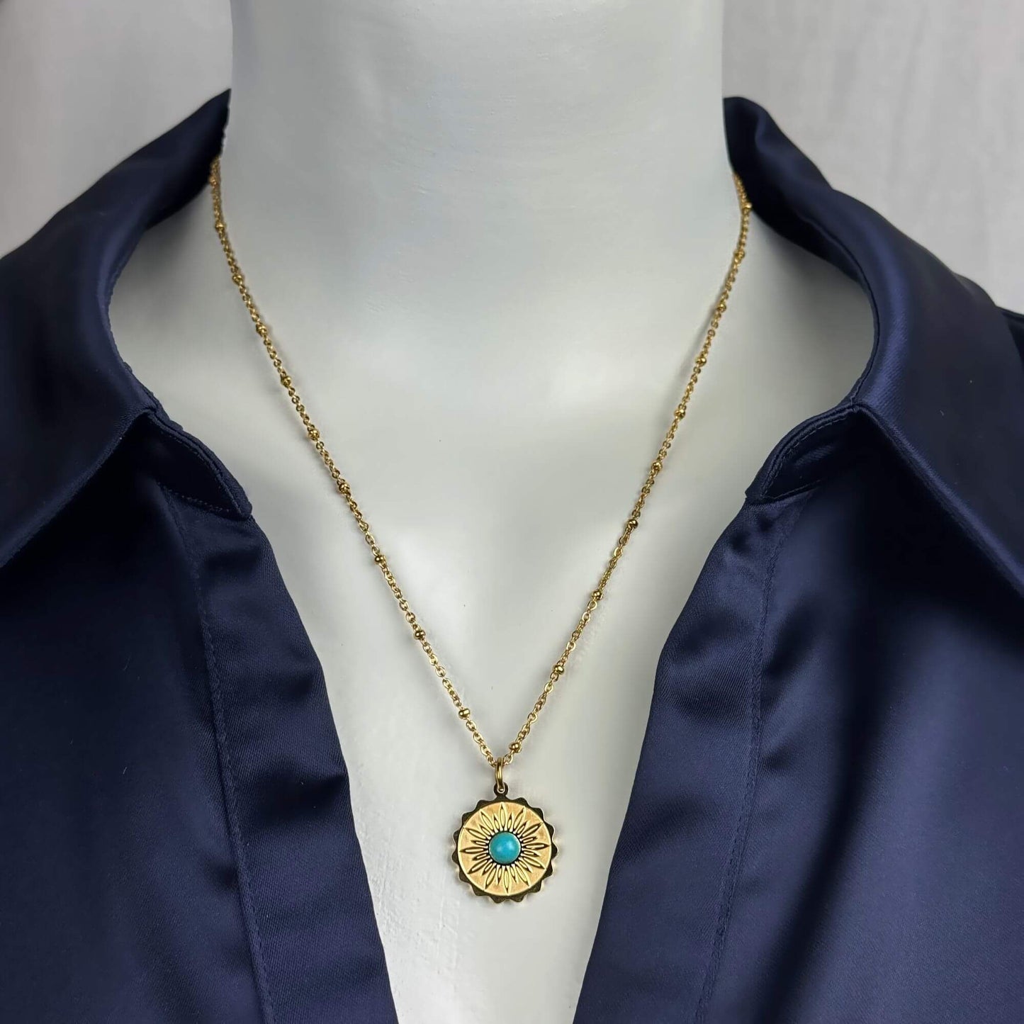 A gold chain necklace with a circular sunburst pendant featuring a turquoise center, displayed on a white mannequin torso wearing a navy satin shirt.