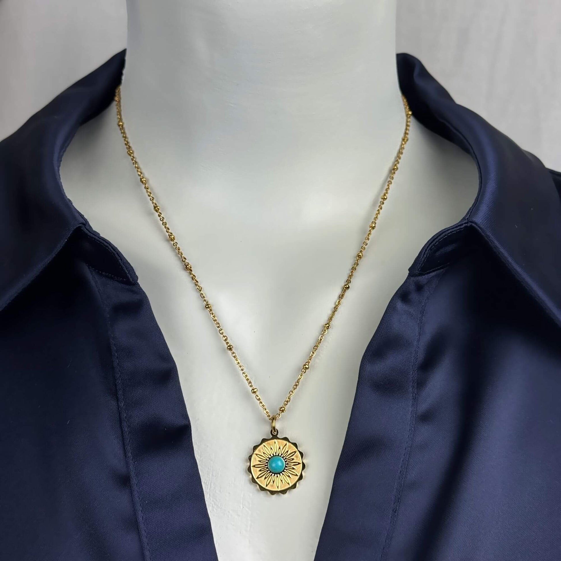 A gold chain necklace with a circular sunburst pendant featuring a turquoise center, displayed on a white mannequin torso wearing a navy satin shirt.