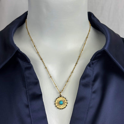 A gold chain necklace with a circular sunburst pendant featuring a turquoise center, displayed on a white mannequin torso wearing a navy satin shirt.