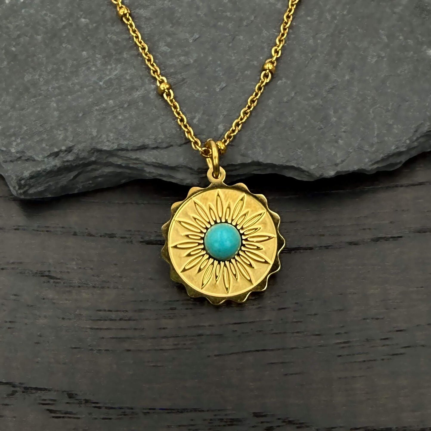 A gold pendant necklace with a sunburst design and a central turquoise stone, displayed on a dark textured surface with a gray slate background.