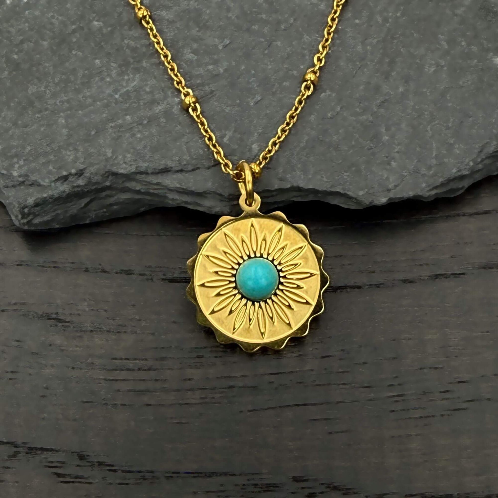A gold pendant necklace with a sunburst design and a central turquoise stone, displayed on a dark textured surface with a gray slate background.