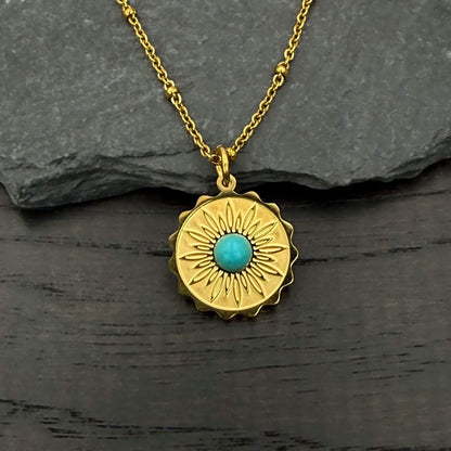 A gold pendant necklace with a sunburst design and a central turquoise stone, displayed on a dark textured surface with a gray slate background.